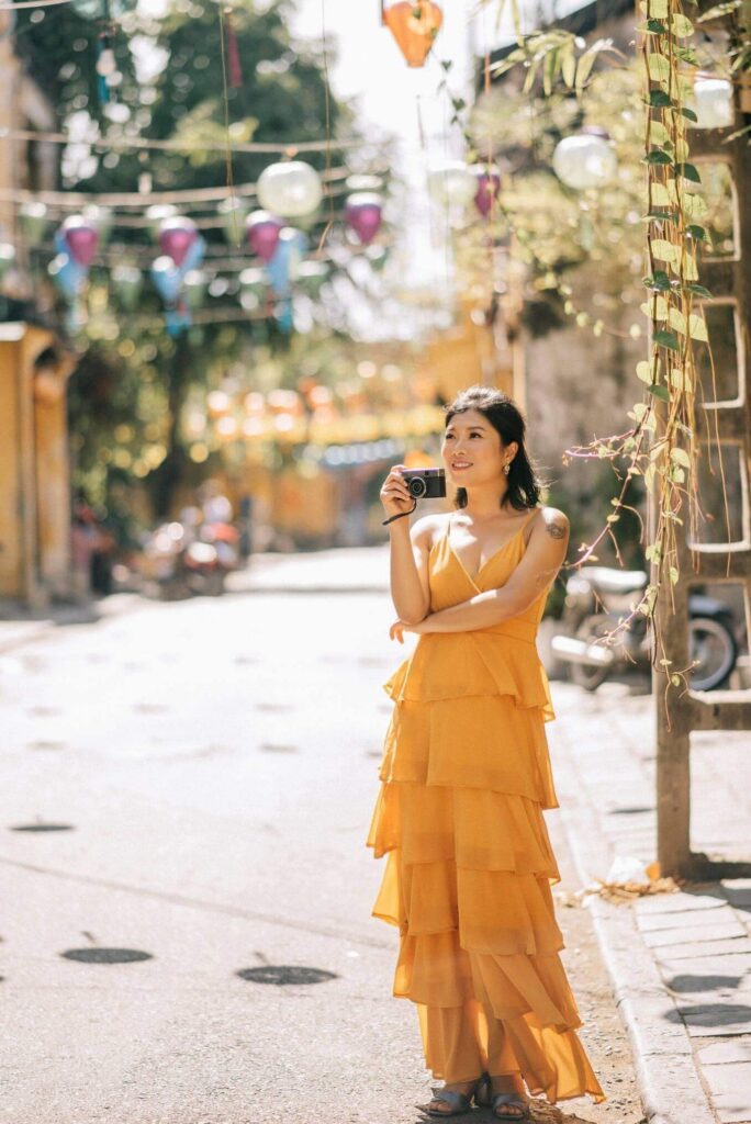 Portrait of a smiling woman in sunlight holding a camera, standing in the colorful streets of Hoi An, Vietnam